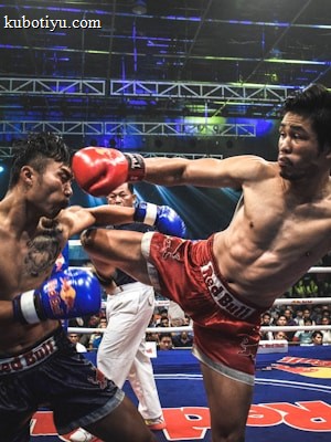 Two athletes are engaged in a dynamic kickboxing match inside a ring, surrounded by a cheering audience. One fighter, wearing red gloves and shorts, executes a high kick towards his opponent, who attempts to block the attack with his arm. The background features colorful lighting and promotional banners.