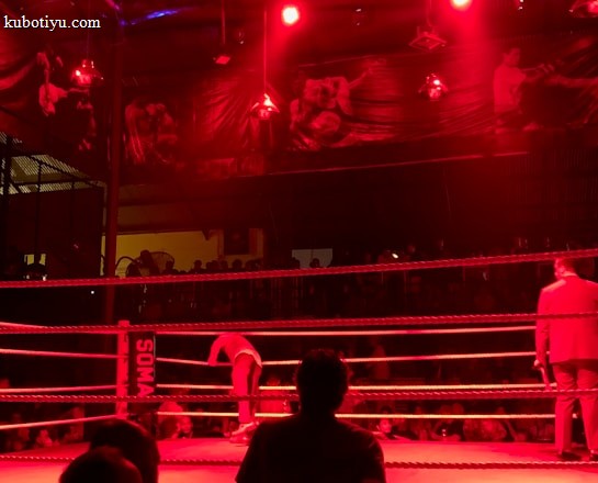 A wrestling ring is illuminated with red lighting, creating a dramatic atmosphere. Several people are visible around the ring, with silhouetted spectators in the foreground. Banners or art adorn the upper walls, and the venue has an industrial ceiling with exposed beams.